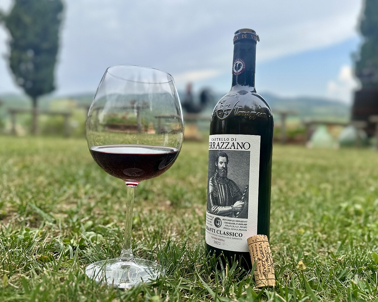芝生の上に置かれた赤ワインの瓶とグラス。瓶の首に黒い雄鶏の丸い印があり、ラベルに CHIANTI CLASSICO と 2022 の文字
