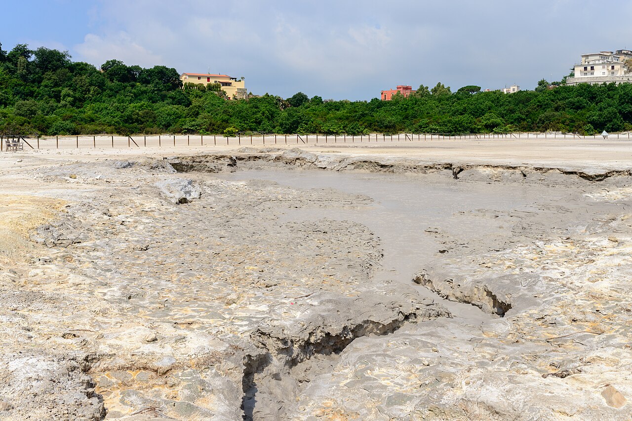 ソルファターラの火口の底。灰色の地面がひび割れ、泥の池が広がる。奥は木々と建物